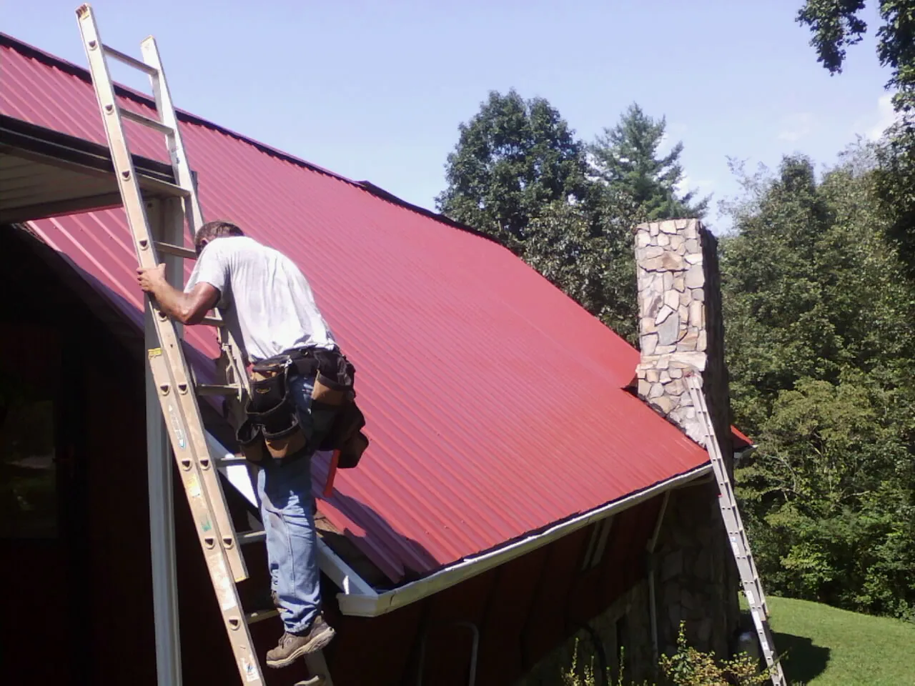 Roofer on a ladder beside a red metal roof and stone chimney — Soffit & Fascia Repair by Roofing Brothers in Hayesville